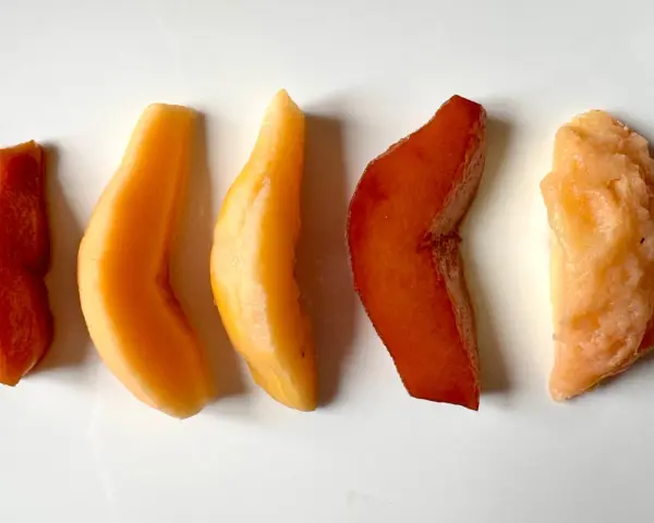 Cinq morceaux de fruit coing cuit, allant du rouge profond à l'orange clair, disposés en ligne sur fond blanc.