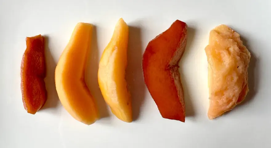 Cinq morceaux de fruit coing cuit, allant du rouge profond à l'orange clair, disposés en ligne sur fond blanc.