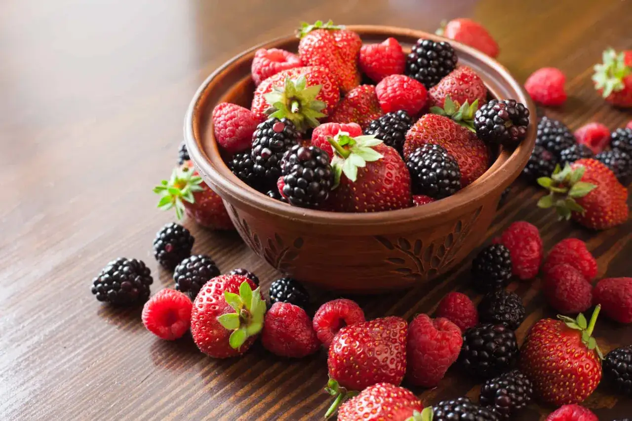 Un bol en terre cuite déborde de fraises, mûres et framboisiers remontants. Quelques fruits sont éparpillés sur la table en bois.